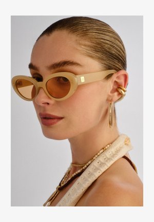 Femme aux cheveux tirés en arrière portant des lunettes ovales beiges, de grandes boucles d'oreilles en or, des colliers superposés de perles et un haut crème texturé.