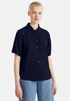 Persona con cabello rubio corto que lleva una camisa holgada de manga corta azul marino oscuro abotonada y vaqueros azul claro sobre un fondo liso.