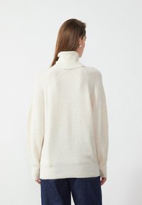 Pull en crème clair, texture côtelée, col montant, épaules tombantes et coupe décontractée. Associé à un pantalon en denim foncé.