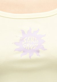 Απαλό κίτρινο ριμπ κορμάκι με ξεθωριασμένο μοβ γραφικό ήλιου και κείμενο "Santa Cruz" στο κέντρο στην περιοχή του στήθους.