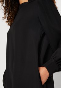 Blusa nera a maniche lunghe con collo alto e polsini arricciati, realizzata in tessuto morbido, con una tasca laterale per un tocco in più.