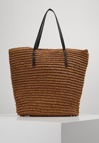 Brązowa torba typu tote wykonana z naturalnych włókien, wyposażona w podwójne czarne skórzane uchwyty, o wyraźnie zarysowanym kształcie i fakturowanej powierzchni.