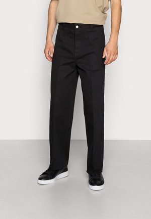 Mężczyzna w czarnych spodniach o prostych nogawkach, beżowej koszuli i czarnych tenisówkach z białymi podeszwami, stoi na szarej podłodze na tle białej ściany.