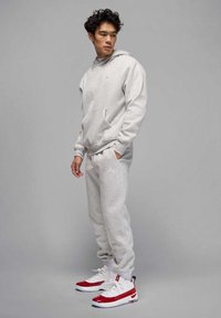 Giovane in felpa grigia chiara e pantaloni della tuta abbinati, con sneakers bianche e rosse, in piedi contro uno sfondo grigio uniforme.
