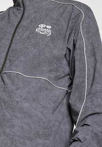 Gray jacket with a textured surface, featuring a white logo, zip closure, and contrasting white piping along the seams.