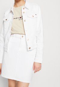 Donna che indossa una giacca di denim bianca e una gonna con bottoni dorati, abbinata a una t-shirt beige con il logo Tommy Hilfiger.