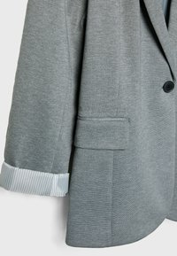Gros plan sur une manche de blazer gris avec une manche retroussée montrant un tissu blanc à fines rayures verticales et une poche à rabat en dessous.