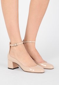 Scarpe con tacco in pelle verniciata beige, con tacco quadrato, punta arrotondata e cinturino alla caviglia con dettaglio in fibbia argentata.