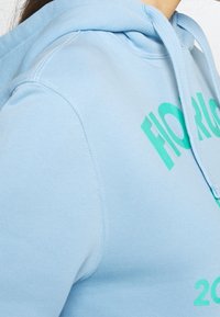 Sudadera con capucha de color azul claro hecha de tela suave con un bolsillo frontal tipo canguro. Cuenta con un diseño de texto en verde y capucha con cordón ajustable.