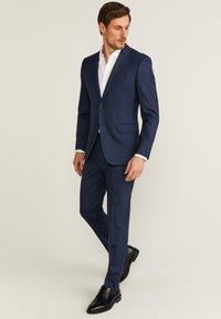 Mörkblå kostym med slim fit, med två knappar och kraaglappar; kombinerad med en vit skjorta och svarta läderskor.