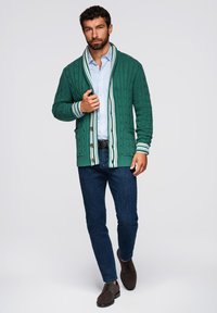 Cárdigan verde de punto trenzado con ribete a rayas blancas y verdes, usado sobre una camisa a rayas azul claro y combinado con jeans oscuros y zapatos marrones.
