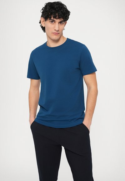 Tricou albastru cu mâneci scurte, realizat dintr-un material moale și neted, cu decolteu clasic rotund. Asortat cu pantaloni închiși la culoare, cu croială lejeră.