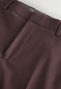 Gros plan sur la ceinture et le pli avant d'un pantalon habillé bordeaux foncé avec passants de ceinture et texture subtile.