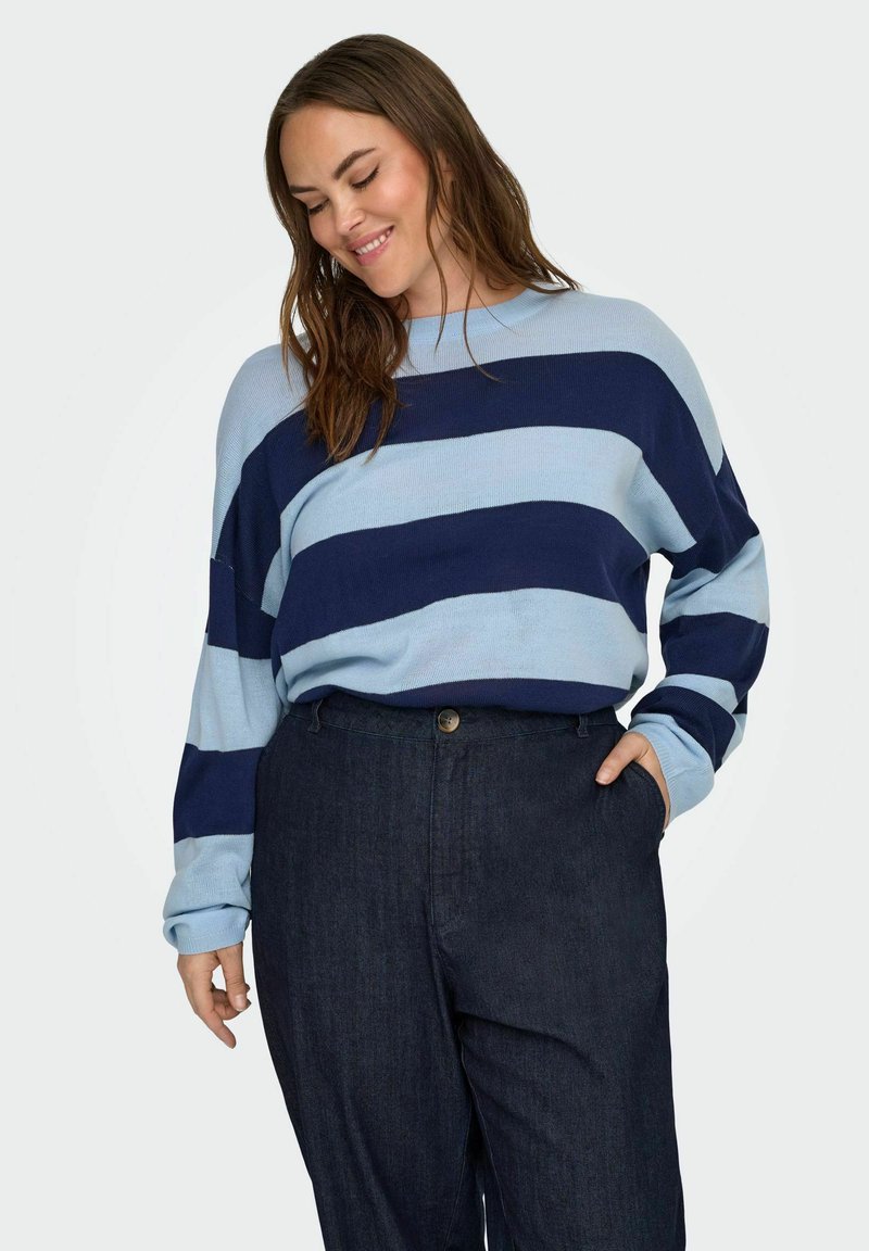 Woman smiling with long brown hair, wearing a light and dark blue striped sweater and dark high-waisted pants, hand in pocket.