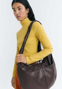 Brown leather shoulder bag with a smooth texture, featuring a wide strap and a subtle seam detail, held by a person wearing a yellow ribbed turtleneck.