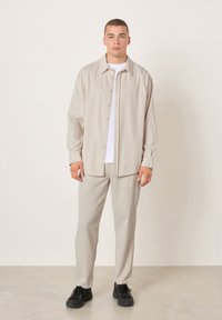 Mladić s kratkom kosom stoji, odjeven u bež rebrastu majicu i hlače s crnim tenisicama, na jednostavnoj svijetloj pozadini.