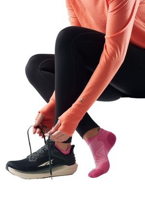 Person iført sorte leggings og koralfarvet langærmet top, der binder sort løbesko over lyserød sportssok på hvid baggrund.