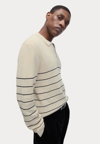 Beige strikket pullover med sorte vandrette striber, der har en krave og en tre-knaps placket. Tekstureret stof, afslappet pasform.
