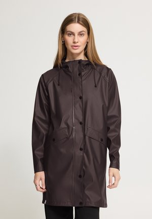 Femme aux cheveux raides portant un long imperméable marron foncé avec des boutons à l'avant et une capuche, se tenant devant un fond clair uni.