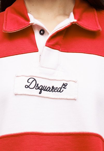 Κοντινό πλάνο ενός κόκκινου και λευκού πουκάμισου με γιακά, με μαύρο κουμπί και το κεντημένο λογότυπο "Dsquared²" στο στήθος.