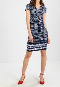 Granatowa dopasowana sukienka z białym abstrakcyjnym wzorem, krótki rękaw i dekolt w serek. W zestawie z czerwonymi szpilkami. Gładka faktura materiału.