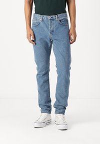 Osoba v světlých modrých džínech a bílých platformových teniskách, stojící na pozadí z čistě bílé barvy.