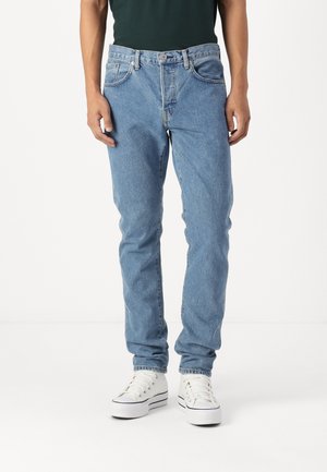 Personne portant un jean en denim bleu clair et des baskets à plateforme blanches, se tenant devant un fond blanc uni.