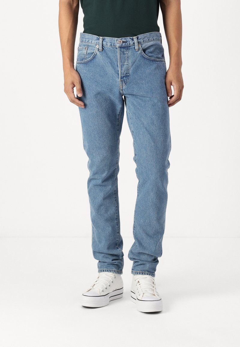 Osoba v světlých modrých džínech a bílých platformových teniskách, stojící na pozadí z čistě bílé barvy.