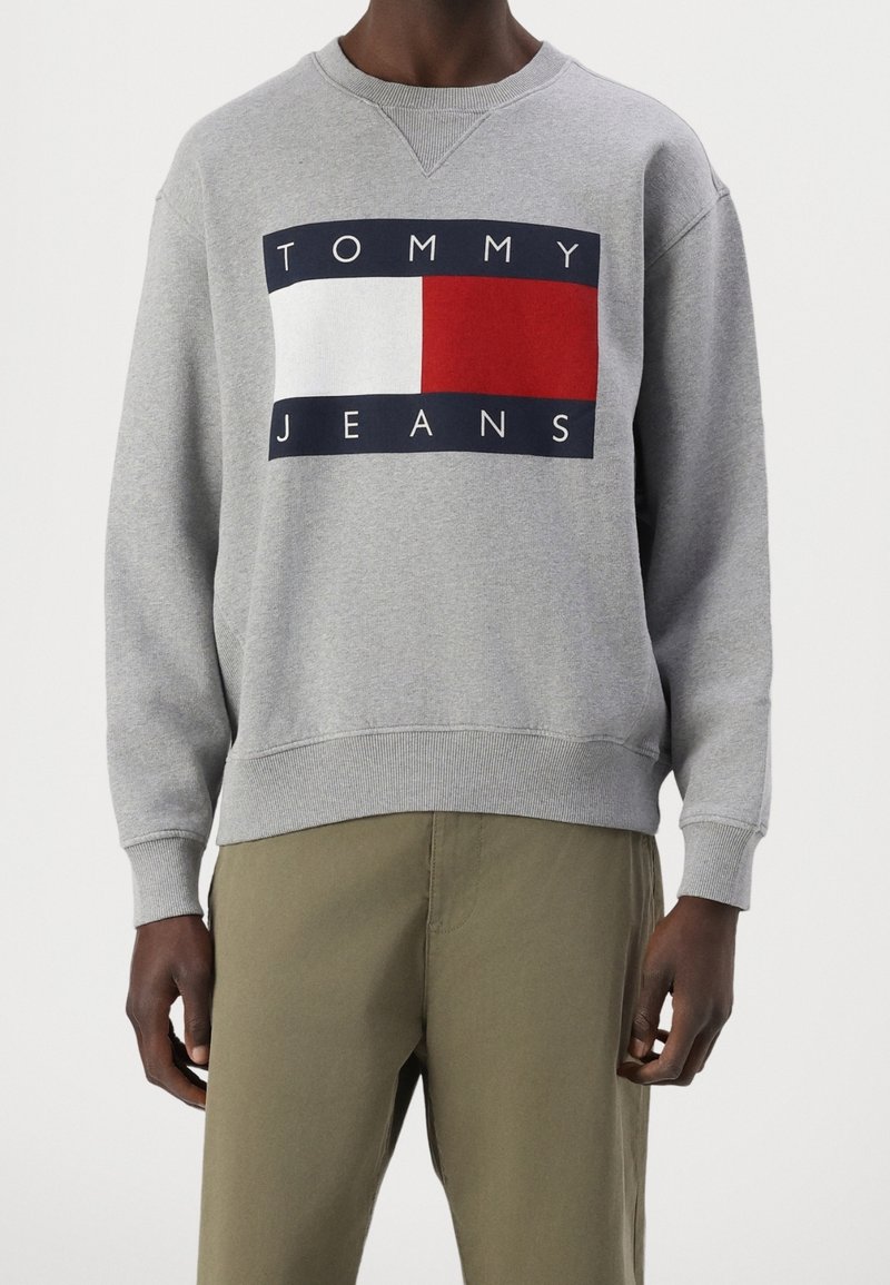 Sweat-shirt gris avec un col rond, arborant un logo Tommy Jeans audacieux en rouge et blanc sur un fond rectangulaire bleu marine. Manches avec poignets côtelés.
