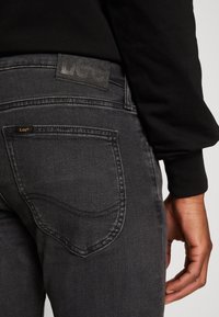 Jean en denim noir à texture lisse, avec détails sur la poche arrière et écusson en cuir du logo Lee au-dessus de la poche.