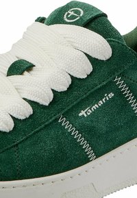 Sneaker verde in scamosciato con lacci bianchi, caratterizzato da un design cucito e un logo. Parte superiore testurizzata e una robusta suola di gomma bianca.