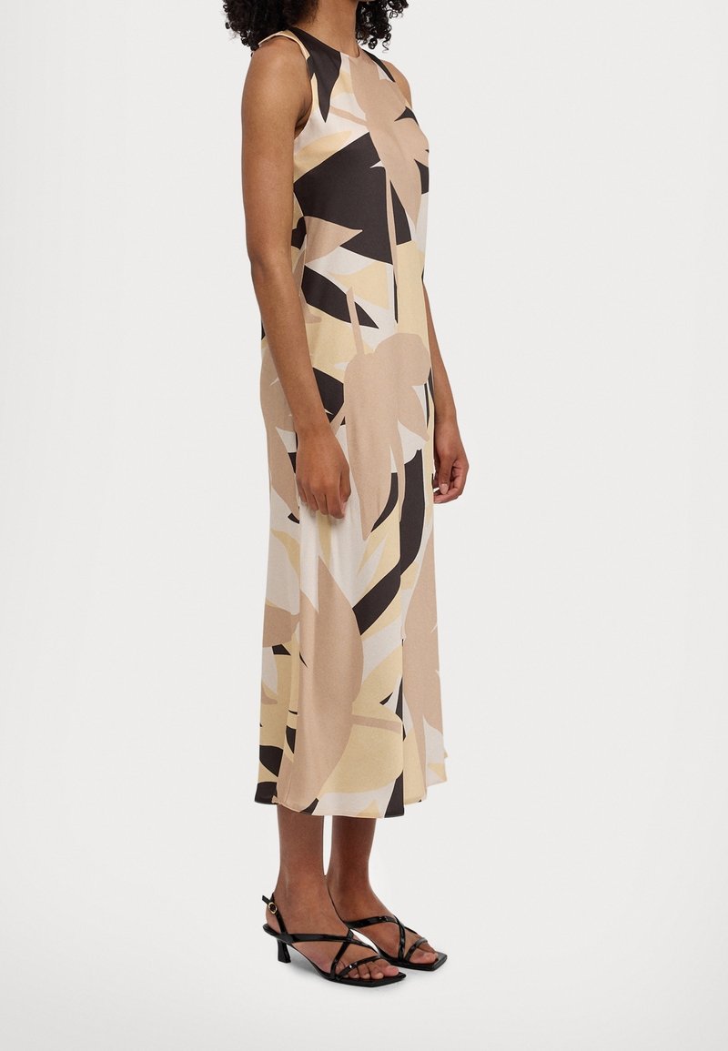 Femme portant une robe midi sans manches avec un motif abstrait beige, noir et blanc et des sandales noires à lanières et talons bas, debout de profil.
