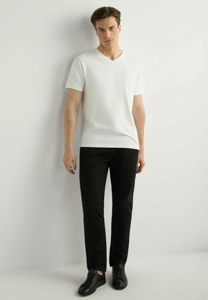 Młody mężczyzna stoi oparty o białą ścianę, ubrany w białą koszulkę z dekoltem w serek, czarne spodnie i czarne trampki, patrząc prosto przed siebie.