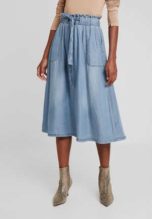 Kvinde iført en lyseblå knælængde denimnederdel med bindebælte, beige langærmet top og støvler ved anklen med slangeskindsmønster.
