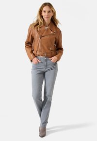 Braune Leder-Motorradjacke mit silbernen Beschlägen, tailliertem Bund und diagonalen Reißverschlüssen, kombiniert mit hellgrauen geraden Jeans.