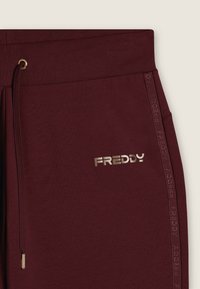 Pantaloni sportivi bordeaux con vita a coste, cordino dorato e accessori in metallo. Presenta un marchio impresso lungo il lato in tessuto coordinato.