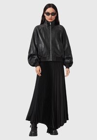 Žena v černé kožené bundě na zip, skládané černé maxi sukni, černých botách s přezkami a úzkých černých slunečních brýlích, stojící na jednoduchém pozadí.