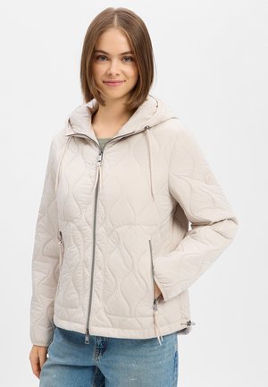 Young woman wearing a light beige quilted zip-up jacket with a hood and blue jeans, standing with one hand in pocket.