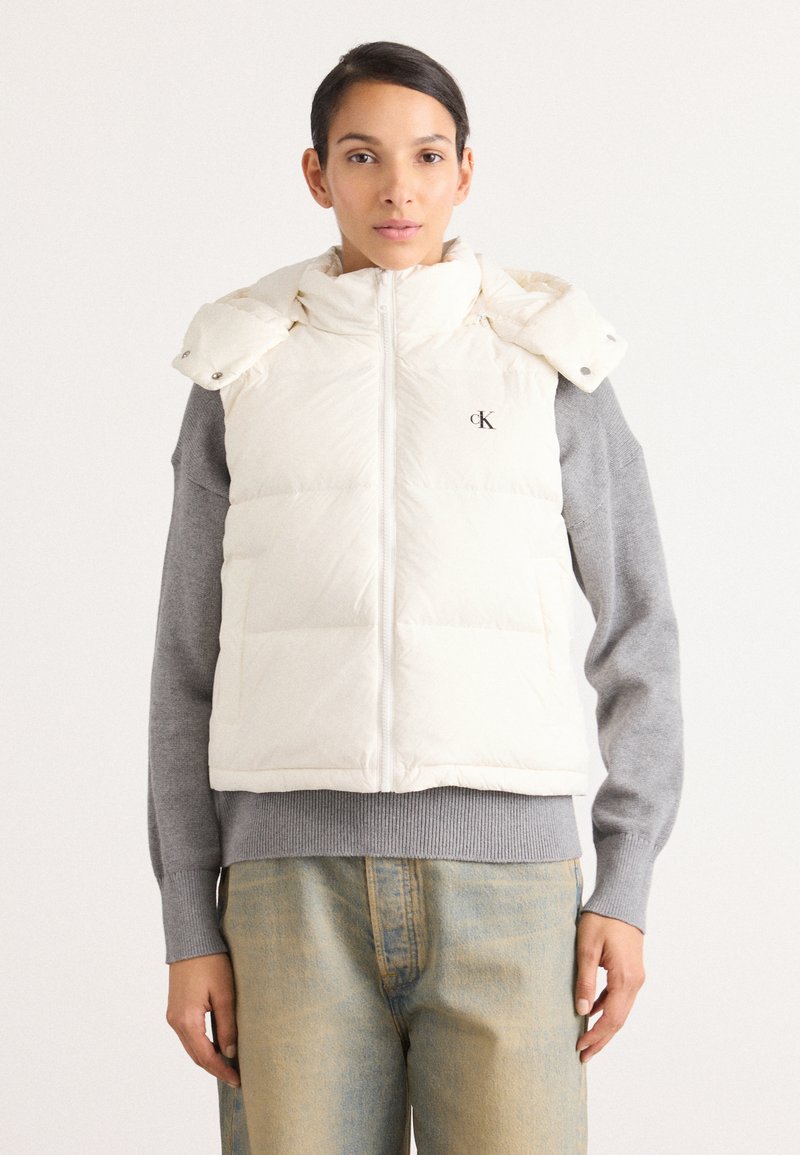 Crème puffer vest met hoge kraag en ritssluiting, voorzien van een logo. Gecombineerd met een lichtgrijze sweater en vervaagde blauwe jeans.