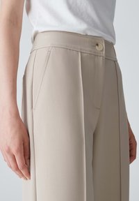 Pantaloni beige su misura con tasche laterali, una piega frontale singola e chiusura con bottone in vita, realizzati in tessuto liscio.