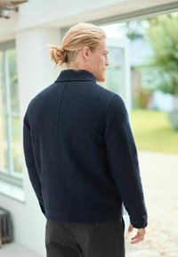 Jachetă din lână de culoare bleumarin, cu guler, având o suprafață texturată și o siluetă ajustată. Detalii cu cusături vizibile la spate și pe mâneci.