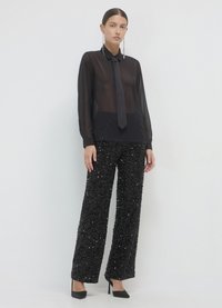 Blouse noire transparente avec col et détail noué, associée à un pantalon large en sequins noirs, et des escarpins noirs. Accessoires minimalistes.
