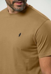 Vīriešu īsām piedurknēm t-krekls gaiši brūnā krāsā, izgatavots no mīkstas auduma. Uz kreisā krūts ir neliels melns izšūts logotips.