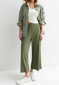 Žena oblečená do olivově zelených širokých ribbed kalhot, bílé tílko, světle olivově zelenou oversized bundu a bílé tenisky, stojící na bílém pozadí.