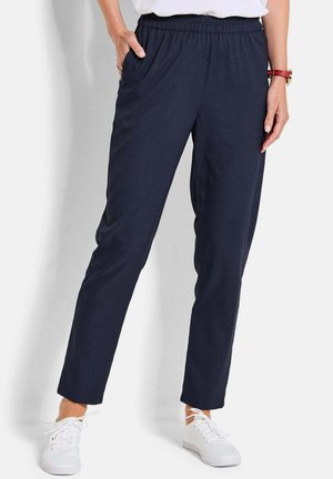 Femme portant un pantalon décontracté bleu marine à taille élastique avec poches latérales, des baskets blanches et un haut blanc, debout avec une main dans la poche.