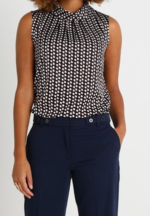 Vrouw draagt een mouwloos zwart-wit topje met patroon, ingestopt in een high-waisted marineblauwe broek, met één hand in de zak en een ring aan de linkerhand.