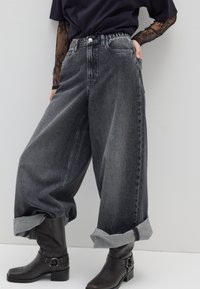 Jeans de mezclilla negros de tiro alto y pierna ancha con una textura deslavada y puños enrollados, combinados con botines negros con detalle de hebilla.