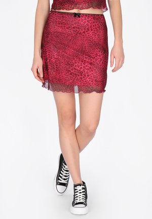 Kobieta w czerwonej spódnicy w cętki lamparta z czarną koronkową lamówką i czarnych wysokich trampkach, stojąca ze skrzyżowanymi nogami.