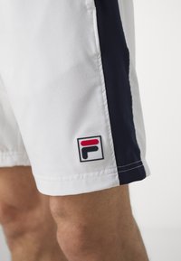 Shorts blancs avec un panneau latéral bleu marine, arborant un logo carré FILA en rouge, bleu marine et noir. Le tissu semble lisse et léger.
