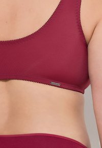 Bordeaux-Bralette mit glatter Textur, breiten Trägern und minimaler Nahtgestaltung. Enthält ein metallisches Logoschild nahe der unteren Kante.